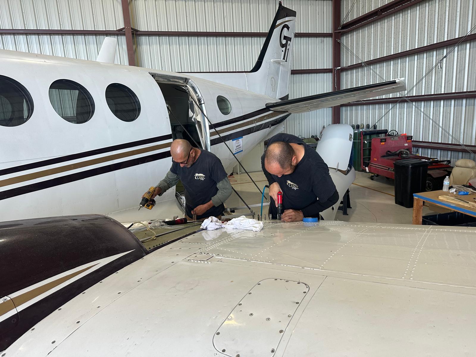 Two 305 Sky technicians working with drills on the wing of a King Air inside the hangar.