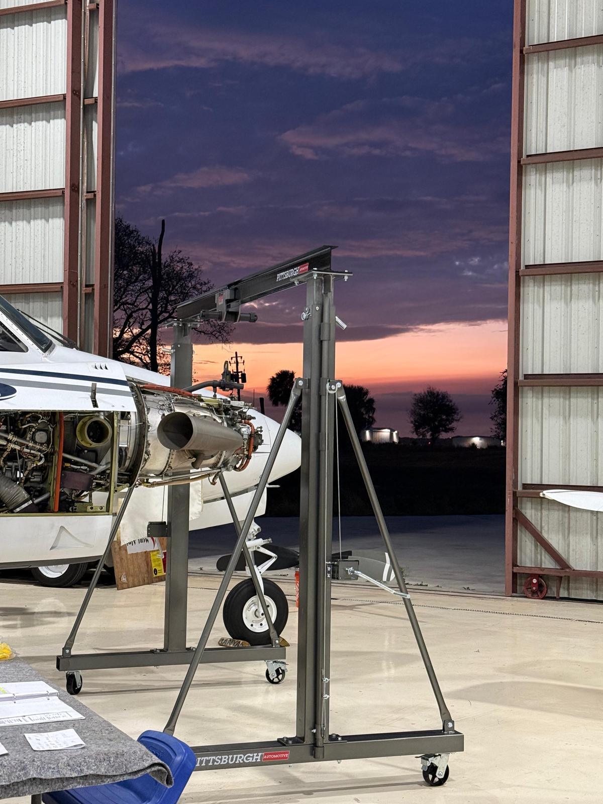 Engine gantry and a King Air with an open nacelle silhouetted against a Florida sunset in the open hangar door.